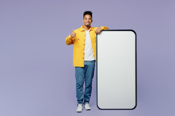 Full body young man of African American ethnicity wear yellow shirt t-shirt big huge blank screen mobile cell phone smartphone with area shwo thumb up isolated on plain pastel light purple background.