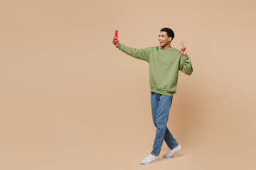 Full body young man of African American ethnicity wear green sweatshirt doing selfie shot on mobile cell phone post photo on social network isolated on plain pastel beige background studio portrait.