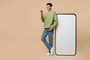 Full body young man of African American ethnicity wear green sweatshirt near big huge blank screen mobile cell phone with area using smartphone device isolated on plain pastel light beige background.