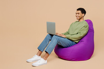 Full body happy young IT man of African American ethnicity wear green sweatshirt sit in bag chair hold use work on laptop pc computer isolated on plain pastel light beige background studio portrait.