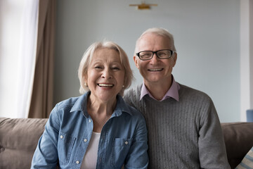 Happy older retired husband and wife sitting on home couch, looking at camera, talking on video call, smiling, laughing, enjoying family conversation. Cheerful elderly couple head shot portrait