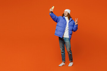 Full body young man with long curly hair wear hat purple ski padded jacket casual clothes doing selfie shot on mobile cell phone post photo on social network isolated on plain orange red background. © ViDi Studio