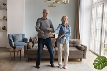 Cheerful energetic senior retired couple celebrating anniversary at home, dancing in living room,...