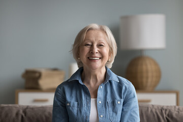 Happy cheerful elderly lady looking at camera with toothy smile, laughing, sitting on couch head...