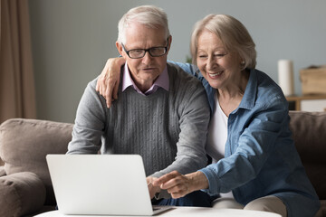 Happy joyful elderly senior couple using online application, service on laptop together shopping, booking hotel for vacation, typing, smiling, laughing, sitting on home couch