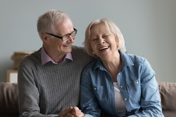 Happy elder husband telling funny story to laughing mature wife. Cheerful old couple sitting on home sofa, holding hands, chatting, talking, having fun, enjoying retirement, marriage anniversary