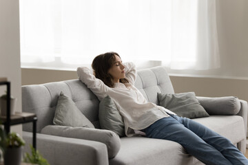 Obraz premium Sleepy tired young adult girl relaxing in home living room, sitting on cozy sofa, resting on couch, sleeping with arms on nap during break, stretching body, enjoying leisure time