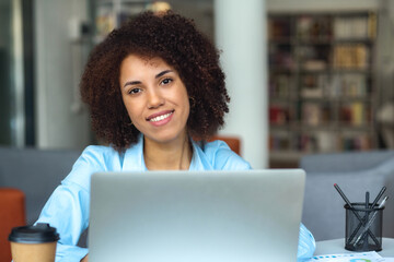 Plakat Portrait of a successful young woman entrepreneur sitting in modern office or at home, working remotely using laptop. Looking at the camera and smiles friendly