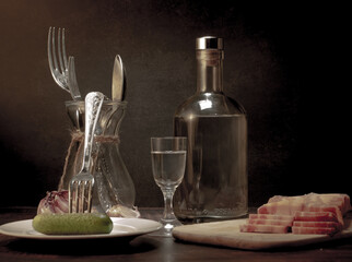 Stylized still life with a bottle of vodka or moonshine with a glass, bacon, sausage and cucumber.