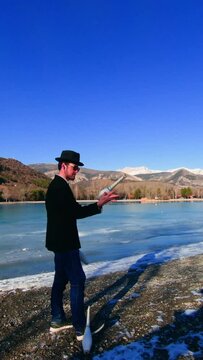 Caucasian Man Juggling 3 White Clubs In Front Of A Frozen Lake. Impressive Fingers Dexterity Trick. He Is Wearing A Formal Jacket With A Deformed Top Hat. Vertical Shot