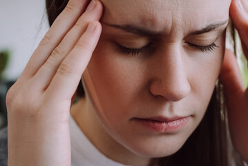 Close up portrait of displeased young woman suffering from migraine while staying alone at apartment. Unhappy millennial lady massaging temples, mental stress, panic attack touching aching sore head
