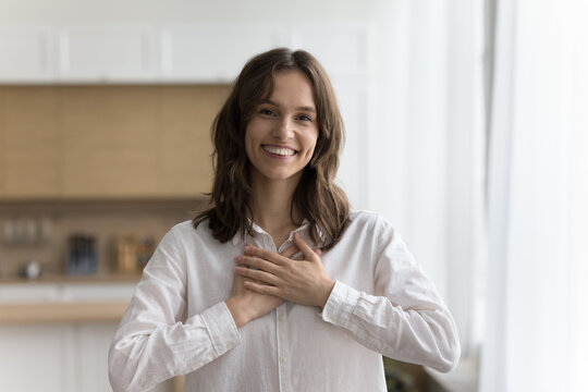 Positive happy young adult girl stacking hands on chest with gratitude, looking at camera, smiling, posing at home. Cheerful woman expressing peace, pride, thank, making friendly gesture of kindness