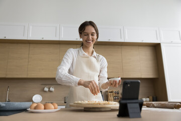 Happy confident young baker woman shooting preparing pizza on cellphone in home kitchen, dropping cheese on dough at smartphone fixed in holder, smiling, laughing