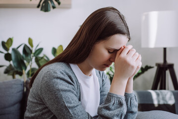 Close up of concerned sad young woman sitting on couch at home thinking of problem looks in...