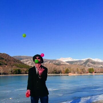man juggling 4 balls (yellow, orange, pink, green) in front of a frozen lake. He wears formal jacket and weird top hat. Mountain in background. Square footage in real time ready for slow motion.