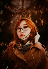 Portrait of a beautiful Asian ginger woman among thin branches with orange leaves in a dark autumn park. Nature in November. Clothing for the season.