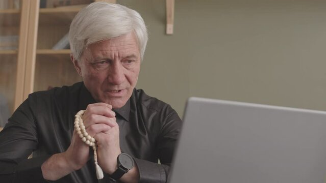 Senior gray haired priest with rosary beads in hands saying prayer during online sermon via laptop, sitting at desk in his home office