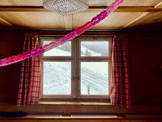 Fenster einer Alphütte, Zeigt in eine schöne verschneite Winterlandschaft in den Bergen.Fenster ist verschlossen. Karierte Vorhänge, rechts und links, rosa Girlande, Holzdecke mit retro Deckenleuchte.