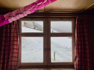 Blick aus dem Fenster. Fenster einer Alphütte, Zeigt in eine schöne verschneite Winterlandschaft in den Bergen.Fenster ist verschlossen. Karierte Vorhänge, rechts und links, rosa Girlande.Holzdecke