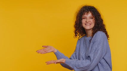 Lovely happy curly haired woman showing thumbs up and pointing empty place, advertising area for commercial text, copy space for goods promotion. Young girl. Studio shot indoors on yellow background