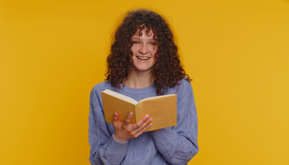 Happy curly haired woman reading funny interesting fairytale story book, leisure hobby, knowledge wisdom, education, learning, study, wow. Young teen girl isolated alone on yellow studio background