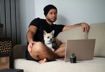 A young, stylish guy in black clothes is at home with his dog. A Chihuahua dog and a guy watch a movie together on a laptop, have fun and spend time together, real friends. The guy and the dog are in 