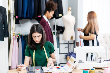 Caucasian female fashion designer works in studio by idea drawing sketches with digital tablet and colorful fabric for a dress design collection, choose clothing colors for tailoring and designing