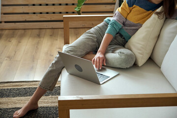 Young woman using laptop, typing, using touchpad, sitting at home on sofa, remote work, work at home