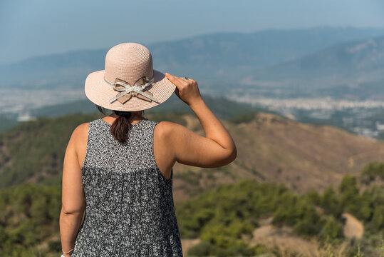Young woman looking into the distance, beautiful mountains in the background, thinking deeply, wearing causal dress and hat, nature, slow life, traveling, brown long hair in a ponny tail
