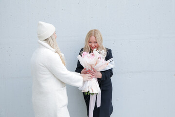 Joyful blonde woman in coat receive gorgeous bouquet of flowers on gray wall. Courier woman hand over order, back view.