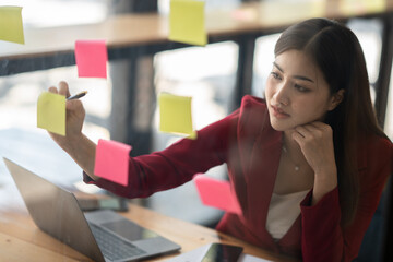 Focused asian business woman writing idea or task on post it sticky notes on glass wall, prepare for the meeting time.