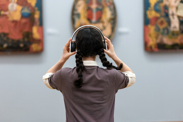 Back view of young woman wearing headphones and contemplates ancient arts. Student visiting gallery or museum. Concept of modern education and culture - obrazy, fototapety, plakaty