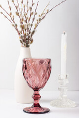 A fluffy willow branch in a vase on the table. A pink wine glass and a white candle . Palm Sunday Celebration