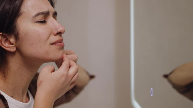 Displeased woman looking in mirror reflection squeezing acnes on skin face in bath room. Focused girl removing pimple on face. Problem skin care