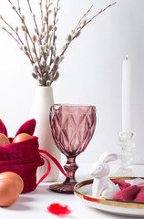 Romantic Easter table setting with candles, white plate, pink wine glass Table decoration with rabbit and painted eggs in red and white colors and a sprig of fluffy willow
