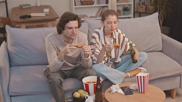 Young Caucasian Couple Staring At TV Screen While Eating Fried Chicken Wings On Couch In Living Room At Night