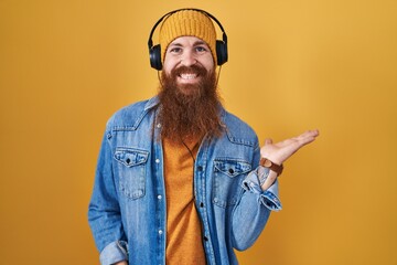 Caucasian man with long beard listening to music using headphones smiling cheerful presenting and pointing with palm of hand looking at the camera.