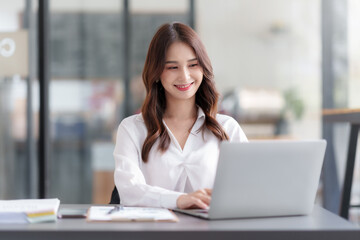 Happy Asian businesswoman sitting in office with her laptop computer working attentively with various documents on her desk.