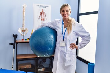 Beautiful woman holding pilates ball at physiotherapy clinic looking positive and happy standing and smiling with a confident smile showing teeth