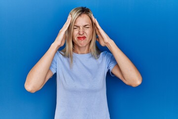 Beautiful blonde woman wearing casual t shirt over blue background with hand on head for pain in head because stress. suffering migraine.