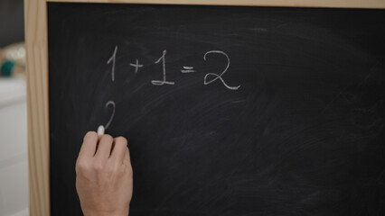 Young blonde woman preschool teacher writing maths exercise on blackboard at kindergarten