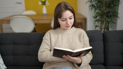 Young blonde woman reading book sitting on sofa at home