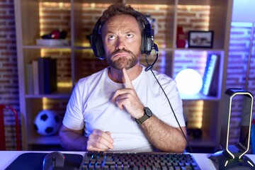 Middle age man with beard playing video games wearing headphones thinking concentrated about doubt with finger on chin and looking up wondering