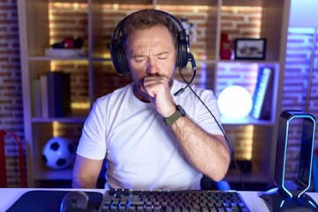 Middle age man with beard playing video games wearing headphones feeling unwell and coughing as symptom for cold or bronchitis. health care concept.