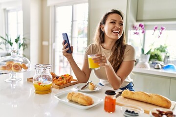 Young beautiful hispanic woman having breakfast using smartphone at the kitchen