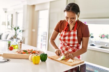 Young beautiful hispanic woman smiling confident cutting onion at the kitchen