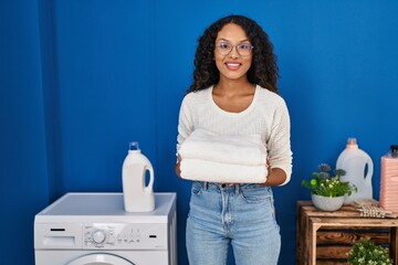 Young latin woman smiling confident holding folded towels at laundru room