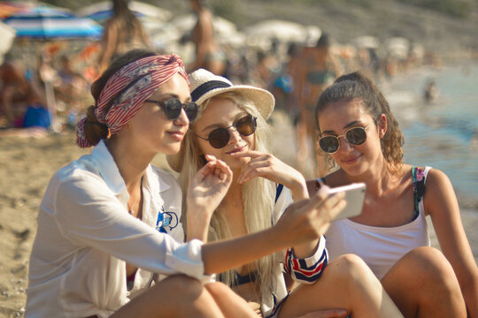 three young women sitting at the beach use a smartphone