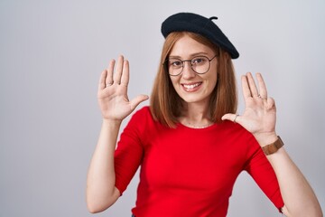 Young redhead woman standing wearing glasses and beret showing and pointing up with fingers number...