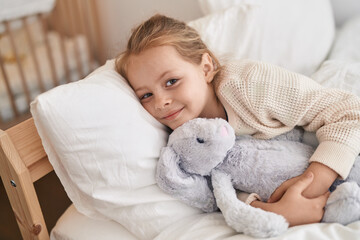 Adorable blonde girl hugging rabbit doll lying on bed at bedroom